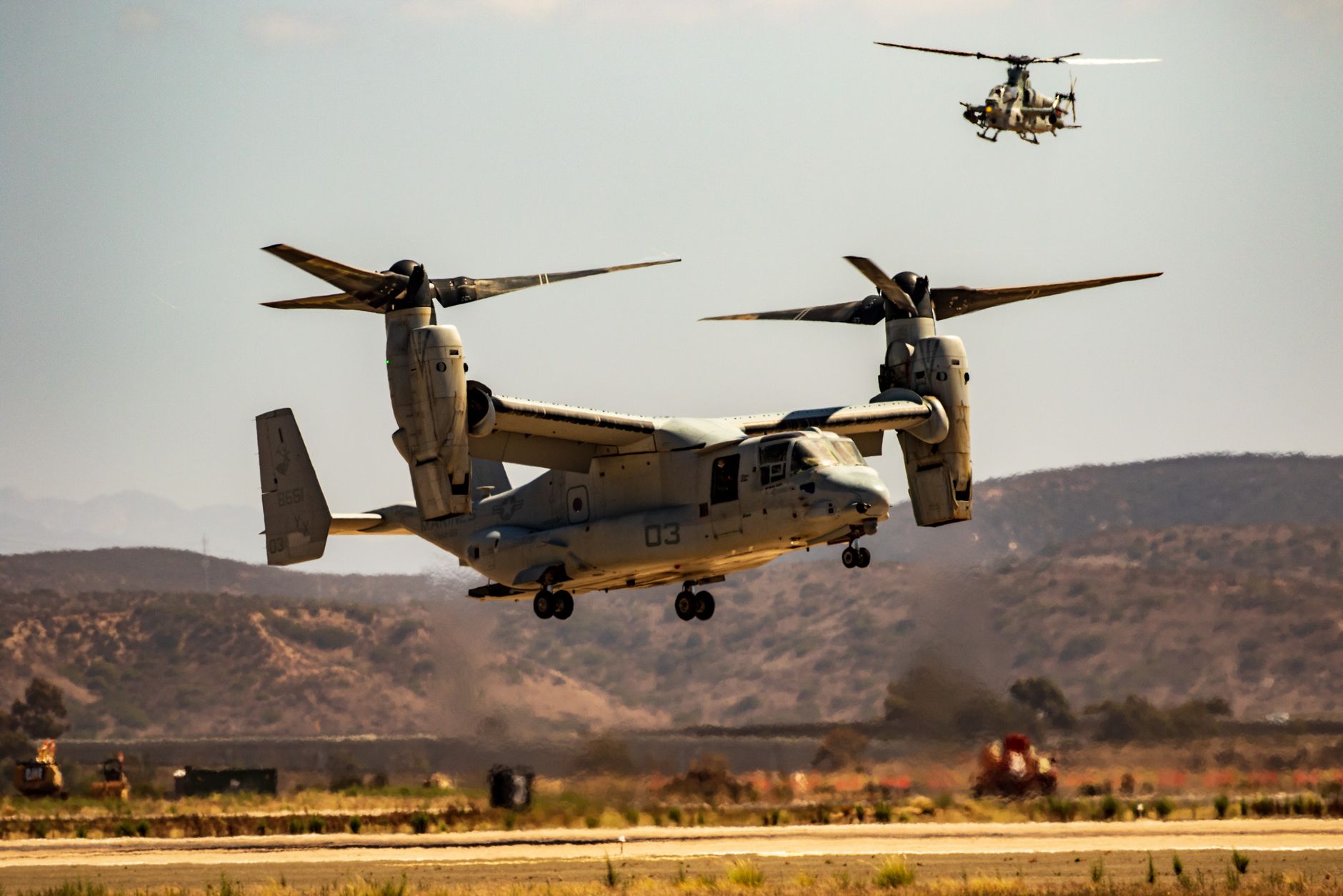 A military Osprey aircraft alongside a helicopter flying over arid landscape in daylight.